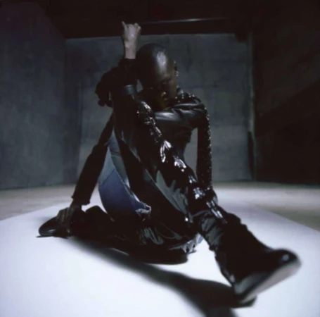 Adult male poses on a white surface in a dark room. He wears a black, glossy jacket and boots, casting a shadow in a dimly lit space.