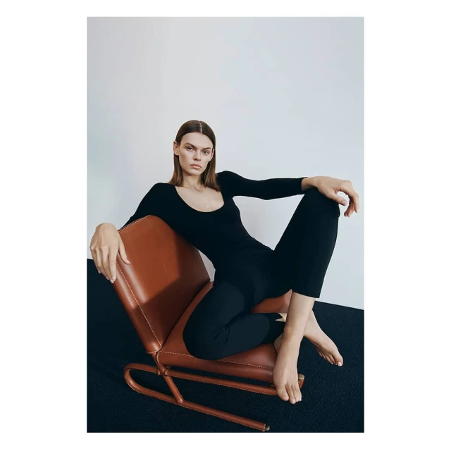 A woman poses in a black long-sleeved jumpsuit, sitting in a modern, tan leather and metal frame chair against a neutral backdrop.