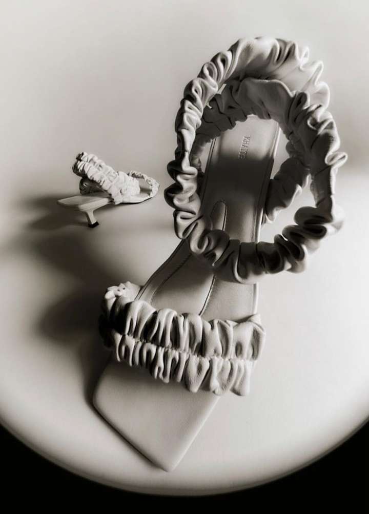 Elegant women's mule sandals with ruched straps on a glossy white surface in a studio setting, creating a high-fashion aesthetic.