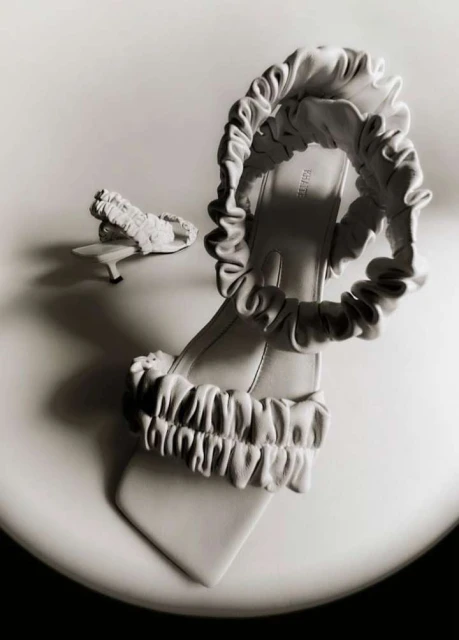 Elegant women's mule sandals with ruched straps on a glossy white surface in a studio setting, creating a high-fashion aesthetic.
