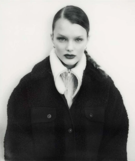 Monochromatic portrait of a woman with slicked-back hair wearing a black, buttoned jacket and a white top, set against a white background.