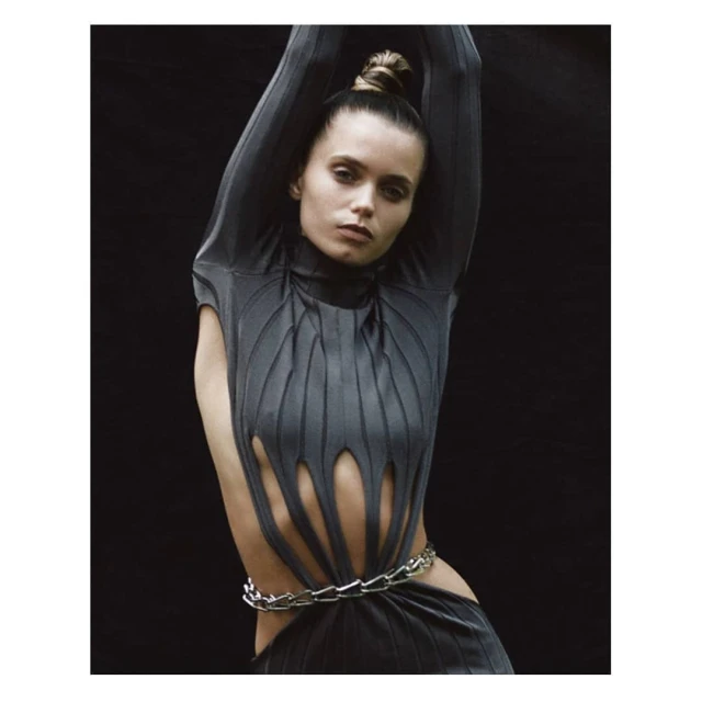Fashion shot of a young adult woman in a gray dress with cutouts, wearing a silver chain belt and her hair in a bun against a black backdrop.