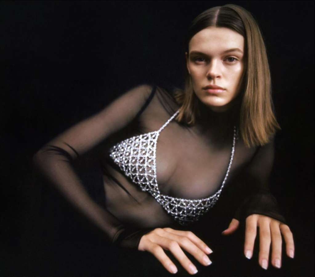 Adult female fashion model poses in a sheer top and beaded bra against a dark background in a studio shot with soft lighting.