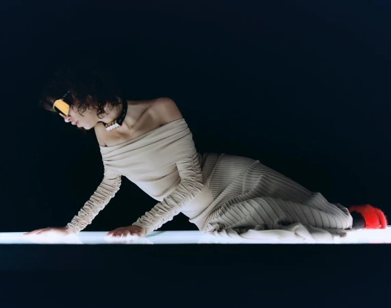 Woman in beige dress and sunglasses poses on a light platform against dark background in a fashion or beauty advertising image.