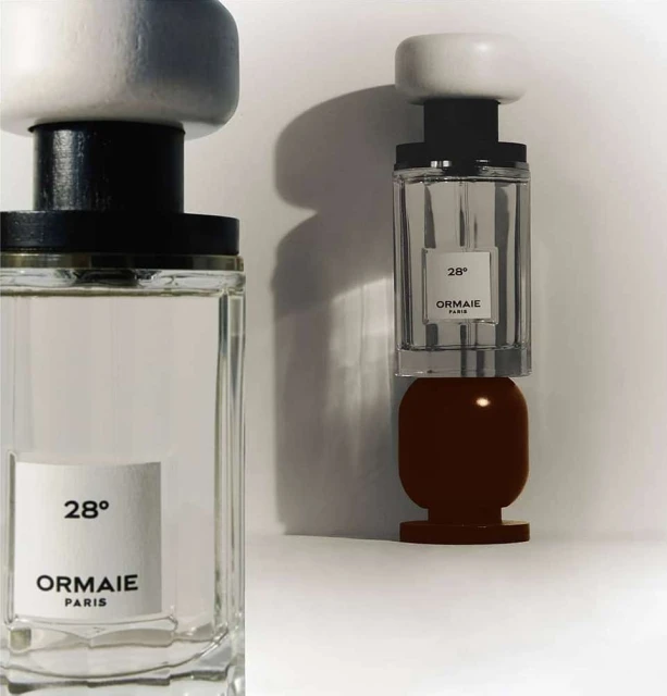 Two Ormaie Paris perfume bottles are displayed, one partially on a dark brown base, against a stark white background and diffused lighting.