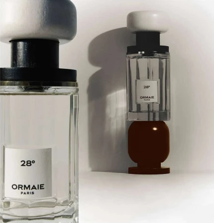 Two Ormaie Paris perfume bottles are displayed, one partially on a dark brown base, against a stark white background and diffused lighting.