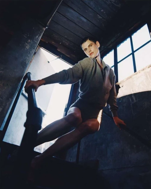 Person in a gray sweater and bodysuit poses dramatically on a wooden staircase with a window, set against a gray interior backdrop.