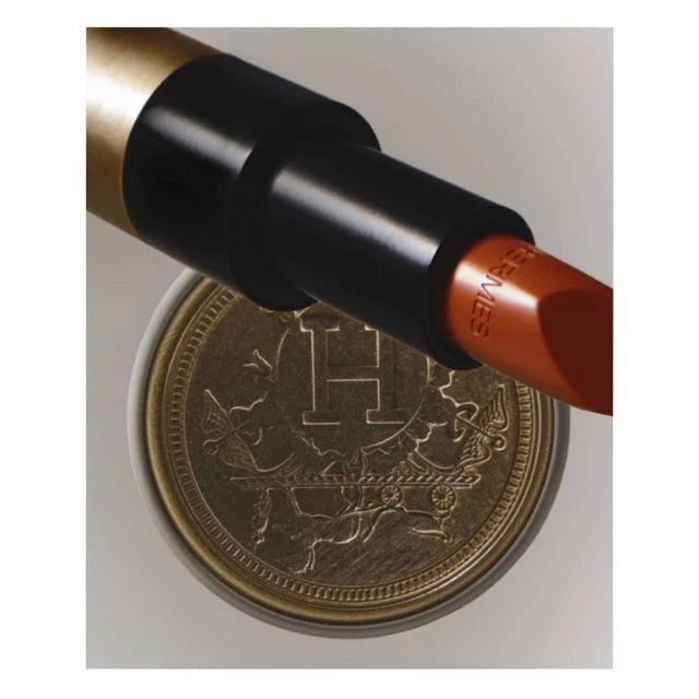 Close-up product shot of Hermes lipstick in a black and gold tube resting on a decorative gold coin with Hermes insignia.