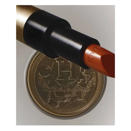 Close-up product shot of Hermes lipstick in a black and gold tube resting on a decorative gold coin with Hermes insignia.