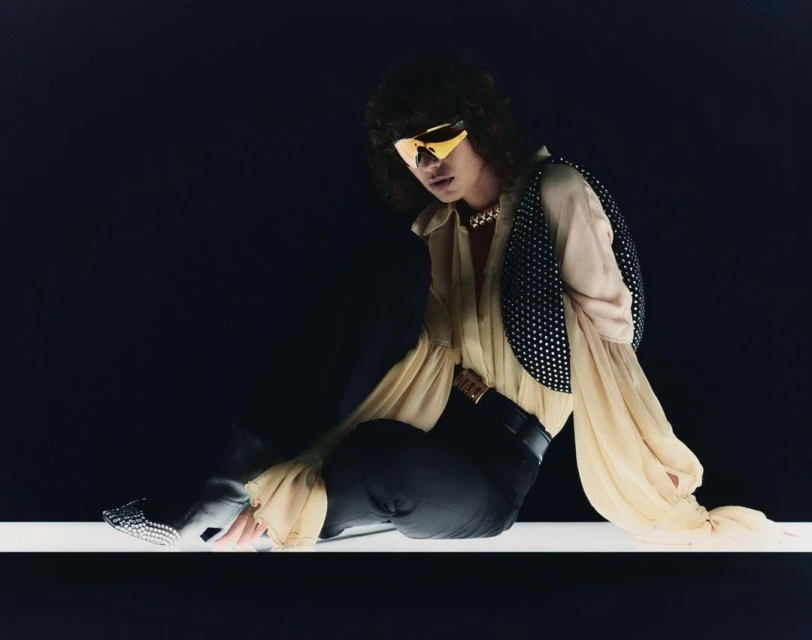 Fashionable individual with sunglasses poses in a studio setting, showcasing a stylish blouse, vest, and dark trousers.