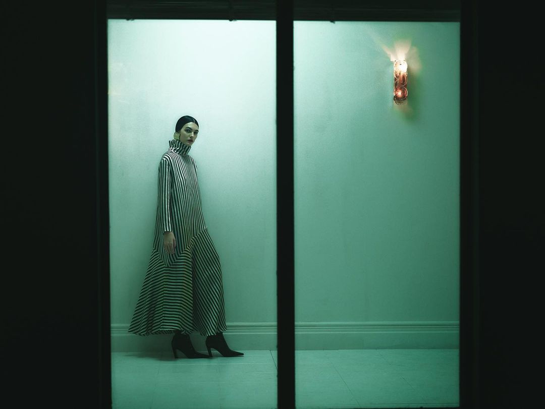 A woman in a black-and-white striped dress stands in a dimly lit hallway, highlighted by an ornamental wall sconce.