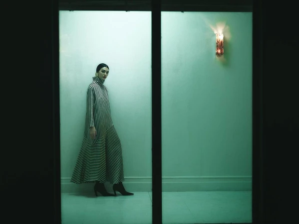 A woman in a black-and-white striped dress stands in a dimly lit hallway, highlighted by an ornamental wall sconce.