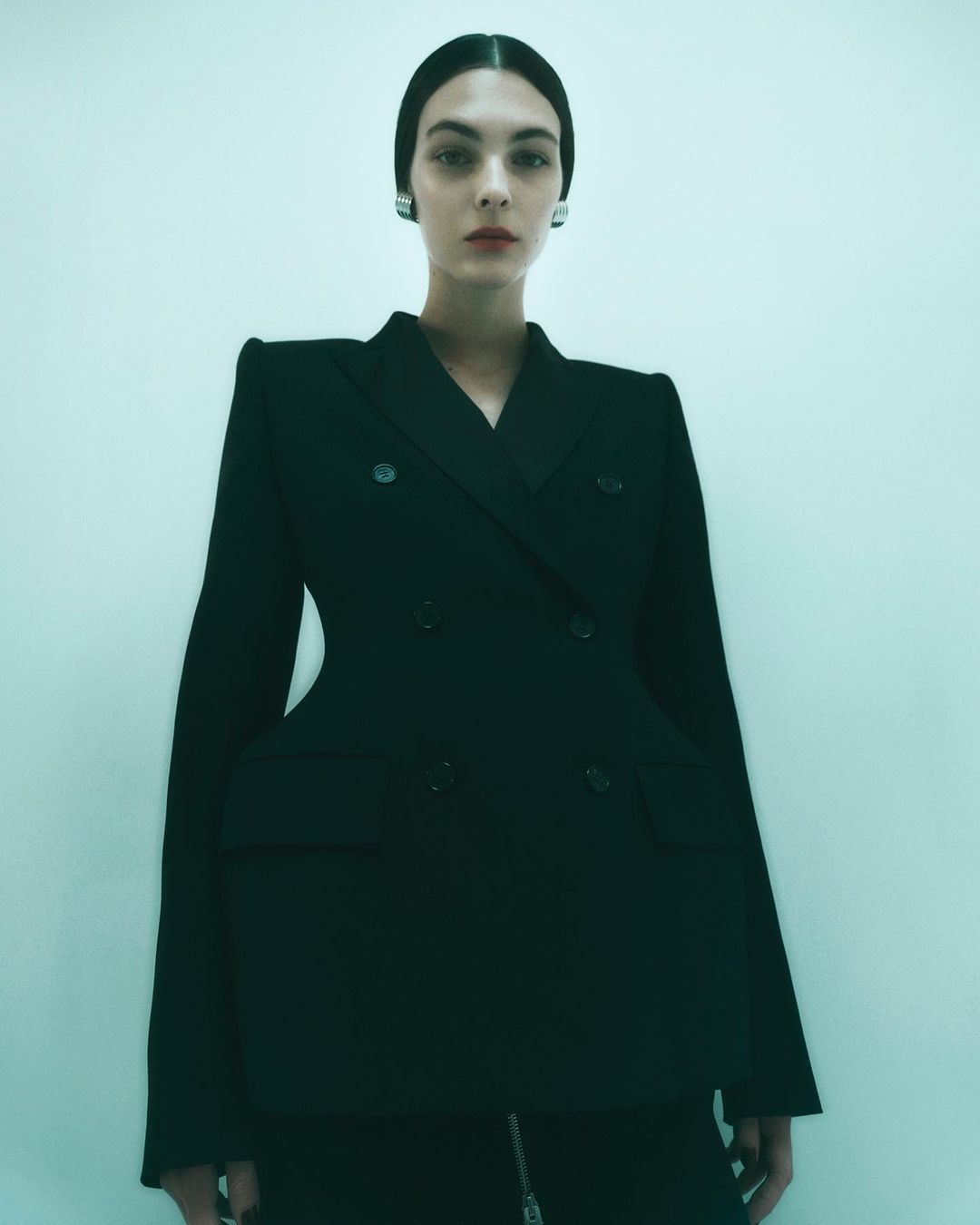 A woman poses in a studio wearing a structured black blazer and skirt, with her hair neatly slicked back, accessorized with silver earrings.