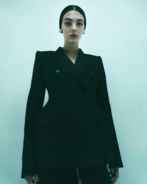 A woman poses in a studio wearing a structured black blazer and skirt, with her hair neatly slicked back, accessorized with silver earrings.