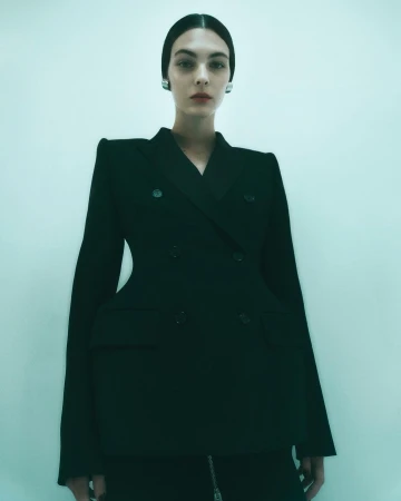 A woman poses in a studio wearing a structured black blazer and skirt, with her hair neatly slicked back, accessorized with silver earrings.