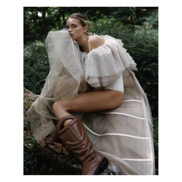 Woman in couture dress and boots posing on a log in a forest. The model has fair skin and hair with a neutral expression.
