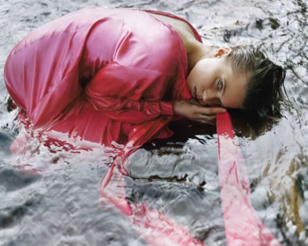 Woman in pink dress lies in water, eyes open, creating a surreal and immersive scene with a blend of fashion and nature.