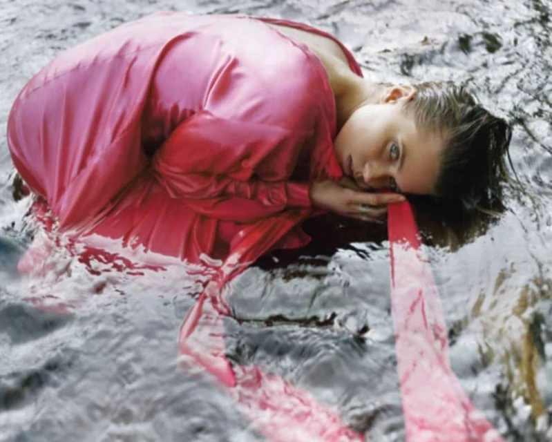 Woman in pink dress lies in water, eyes open, creating a surreal and immersive scene with a blend of fashion and nature.