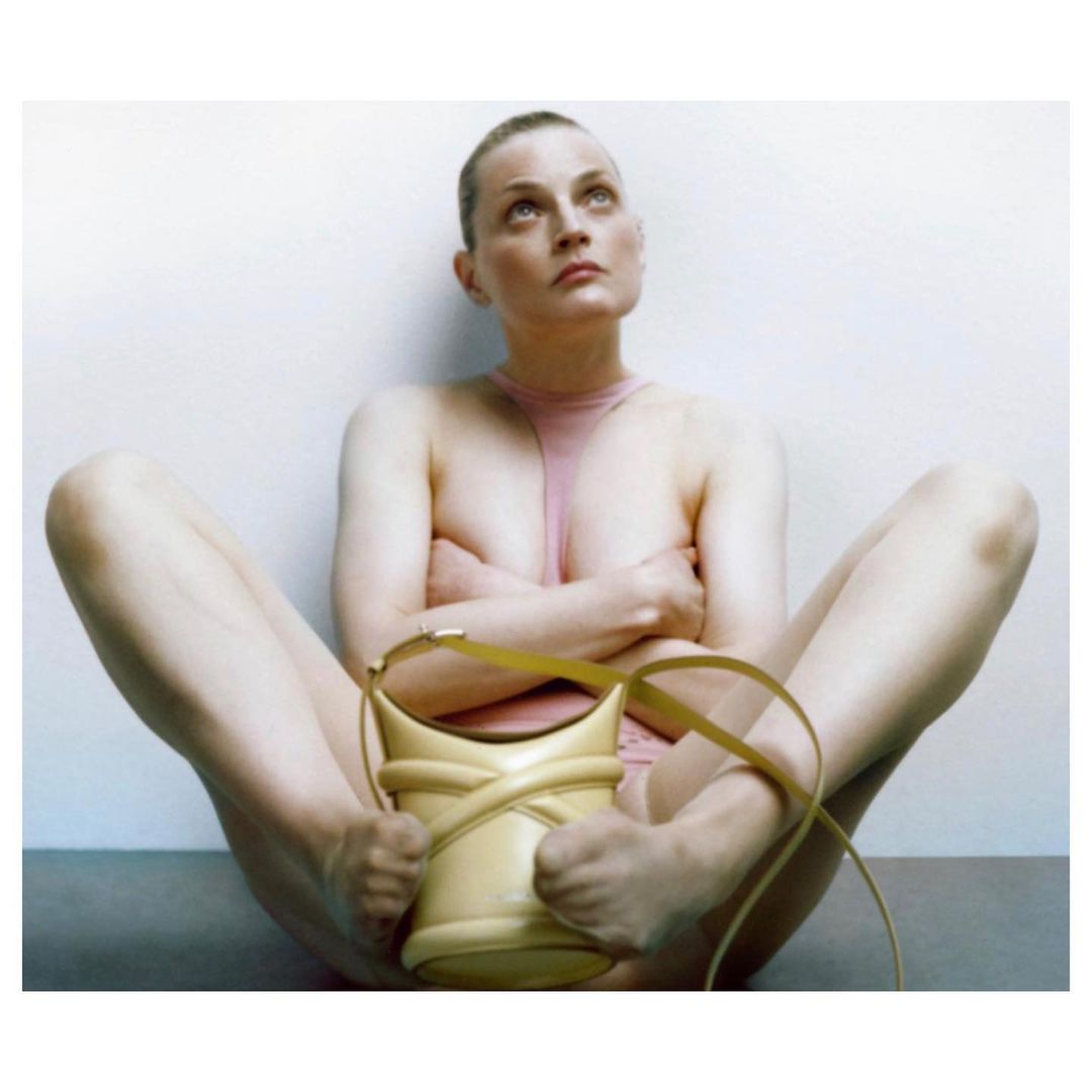 A slender adult woman poses nude indoors, seated on the floor with legs open, hands on her ankles, wearing a light pink top, and handbag.