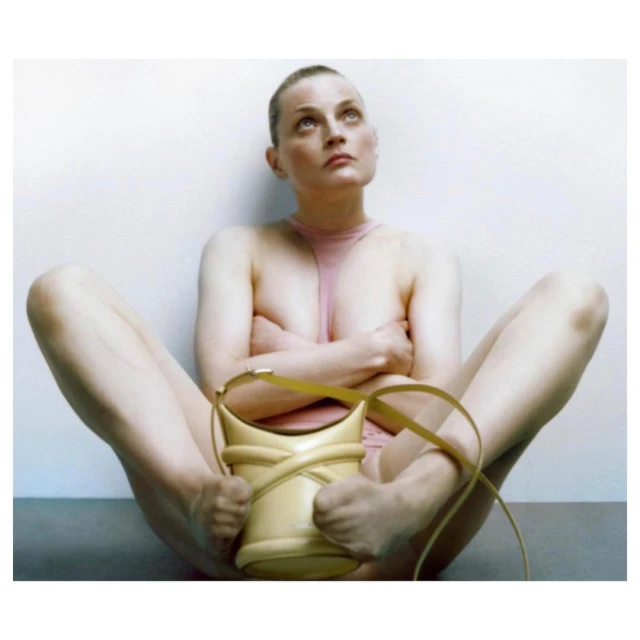 A slender adult woman poses nude indoors, seated on the floor with legs open, hands on her ankles, wearing a light pink top, and handbag.