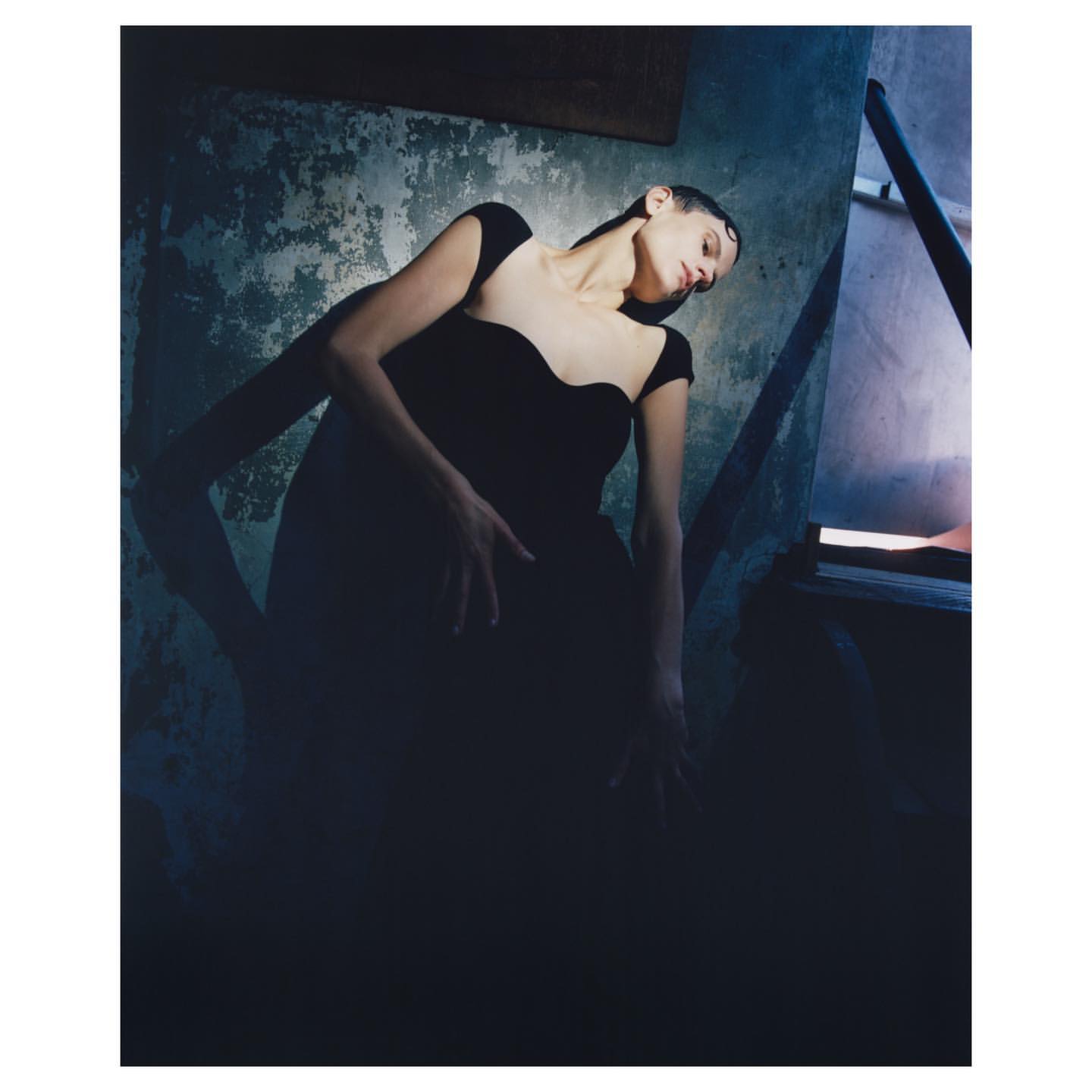 Adult female fashion model poses in black dress near doorway against textured wall in low-key lighting in a moody, artistic image.
