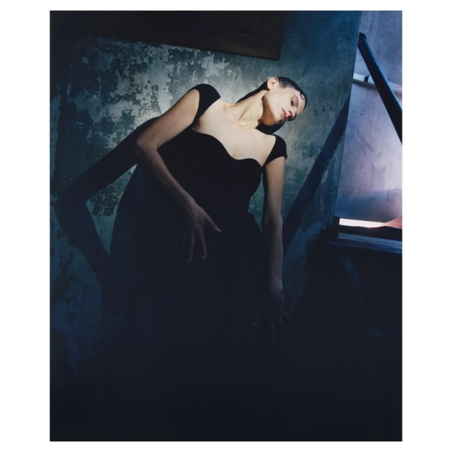 Adult female fashion model poses in black dress near doorway against textured wall in low-key lighting in a moody, artistic image.