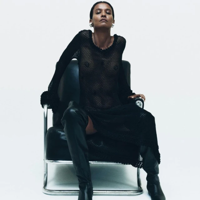 A woman in a black sheer dress sits in a black and silver chair against a white backdrop, creating a minimalist fashion shot.