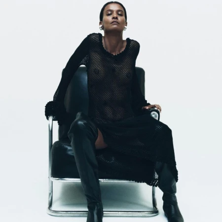 A woman in a black sheer dress sits in a black and silver chair against a white backdrop, creating a minimalist fashion shot.