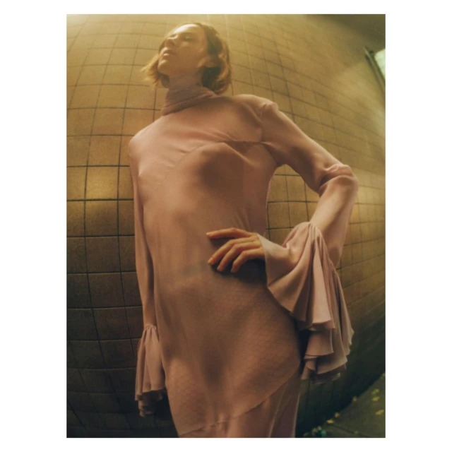 A woman in a pale pink dress with bell sleeves poses against a yellow tiled wall, creating an editorial fashion image indoors.