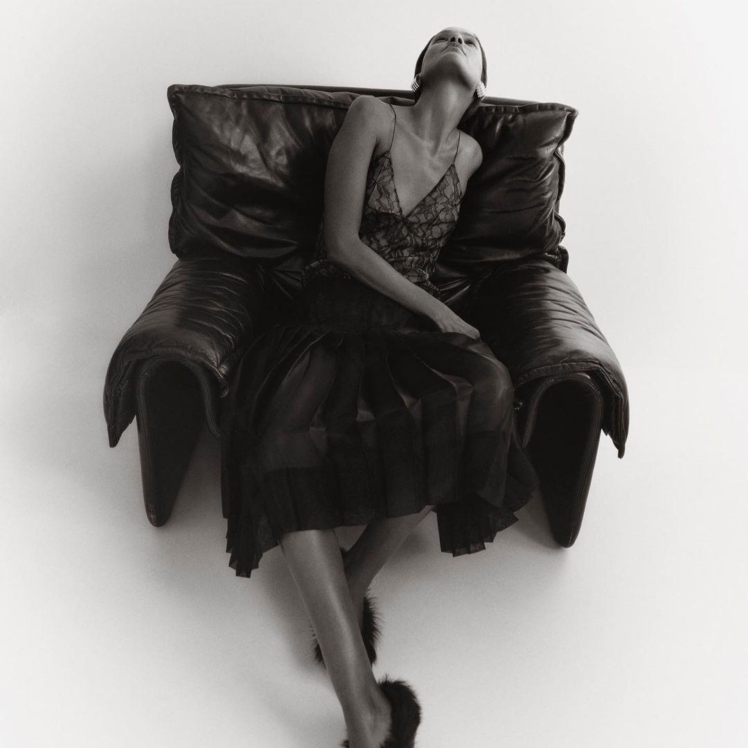 Monochrome portrait of a woman in a slip dress reclining on a large leather chair, captured in a studio setting with diffused light.