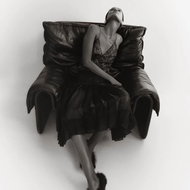 Monochrome portrait of a woman in a slip dress reclining on a large leather chair, captured in a studio setting with diffused light.