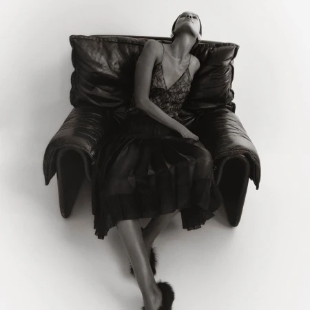 Monochrome portrait of a woman in a slip dress reclining on a large leather chair, captured in a studio setting with diffused light.