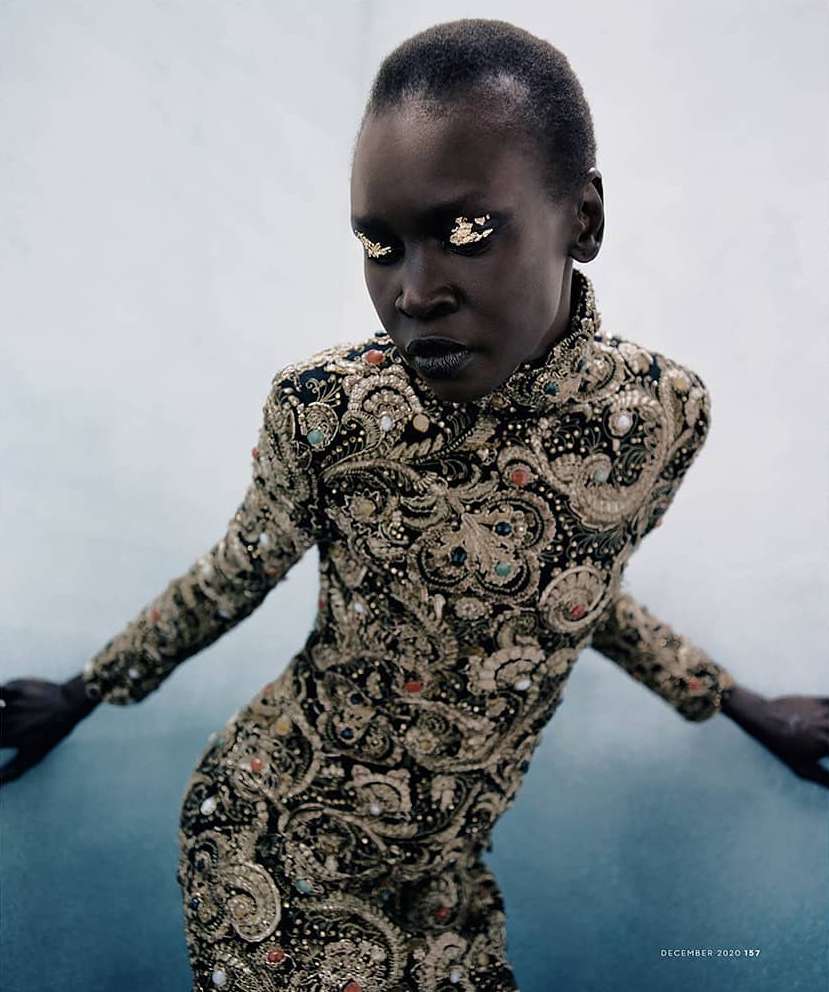 An African female model with golden eyeshadow posing in a black dress with gold embellishments against a gradiated white background.