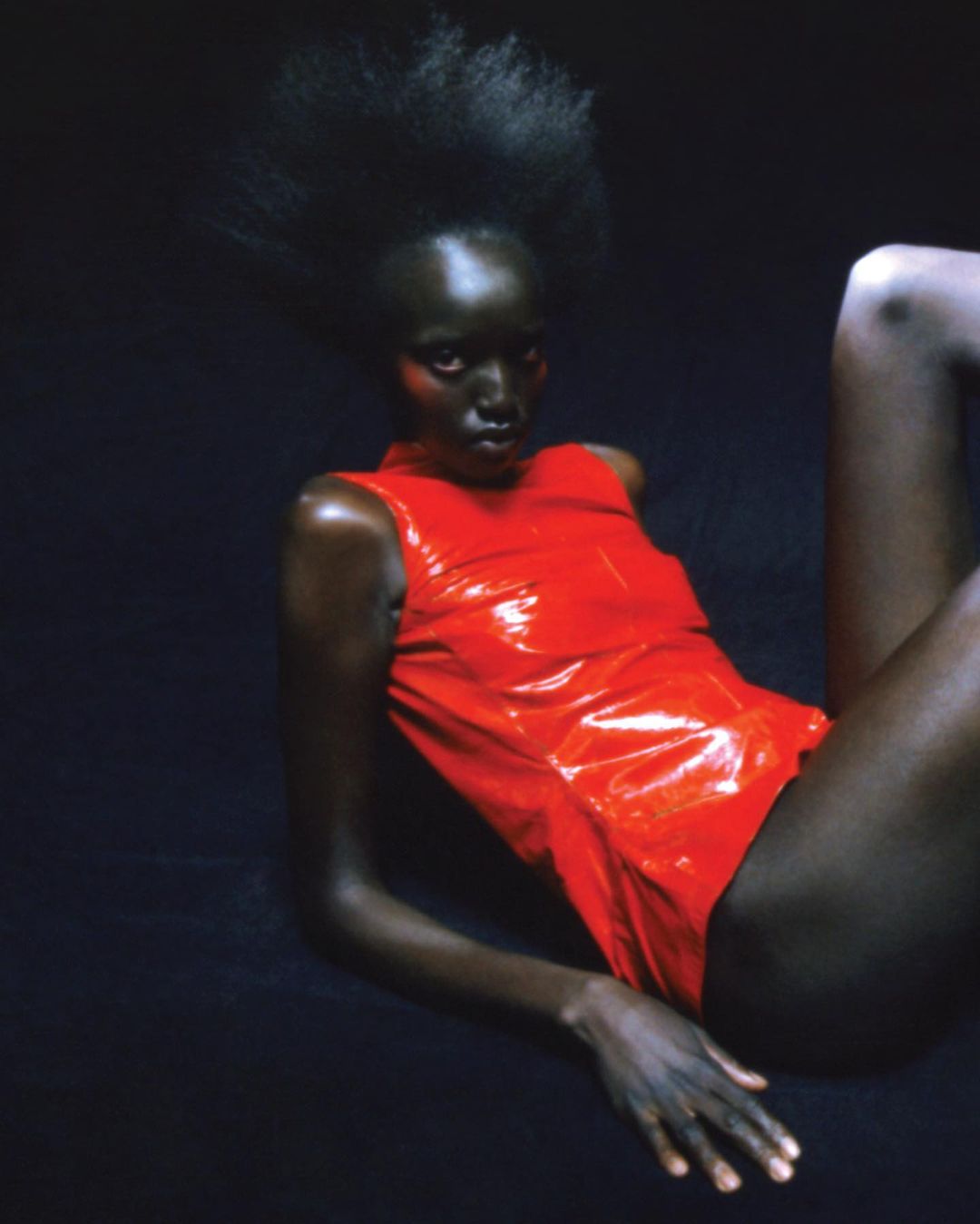 Woman with dark skin and high afro hairstyle poses in shiny red bodysuit against a dark background in studio lighting. Beauty and fashion.