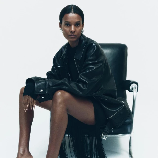 A fashionable woman with dark hair sits in a modern black chair wearing a dark leather jacket, short skirt, and minimal jewelry in a studio setting.