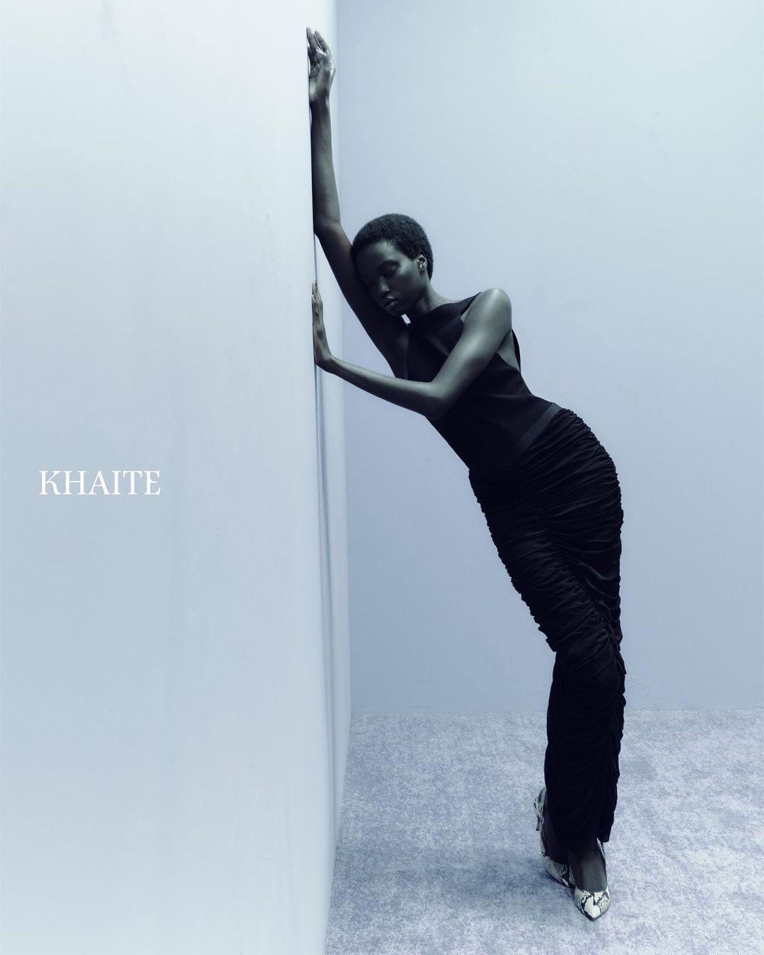 Fashion model poses in a black ruched dress and snakeskin-print heels, leaning against a wall in a softly lit, neutral indoor setting.