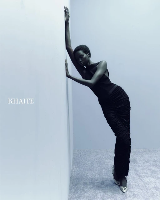 Fashion model poses in a black ruched dress and snakeskin-print heels, leaning against a wall in a softly lit, neutral indoor setting.