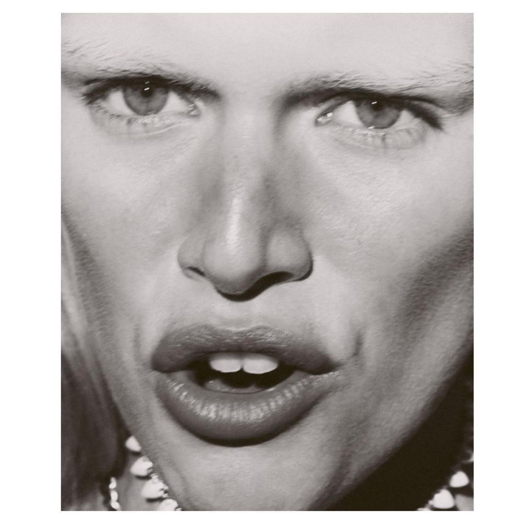Close-up black-and-white portrait of an adult with light skin, pale eyebrows, and a decorative beaded necklace.