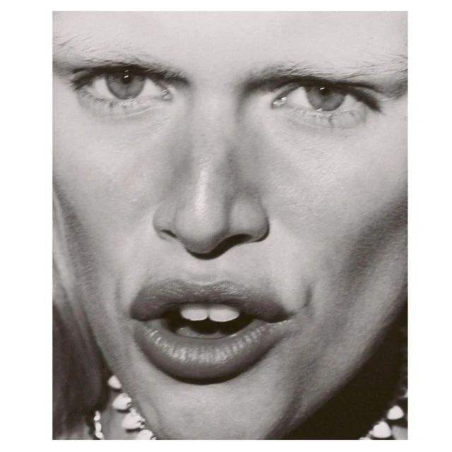 Close-up black-and-white portrait of an adult with light skin, pale eyebrows, and a decorative beaded necklace.