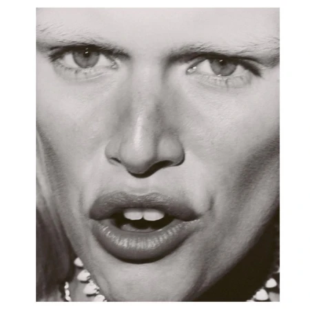 Close-up black-and-white portrait of an adult with light skin, pale eyebrows, and a decorative beaded necklace.