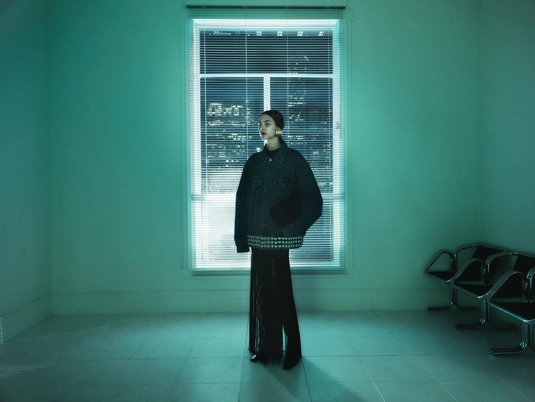 Fashion shot of a woman in a denim jacket and black pants, standing in a room with mint green walls, a window, and metal chairs.