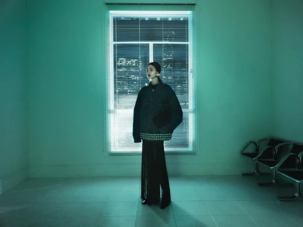 Fashion shot of a woman in a denim jacket and black pants, standing in a room with mint green walls, a window, and metal chairs.