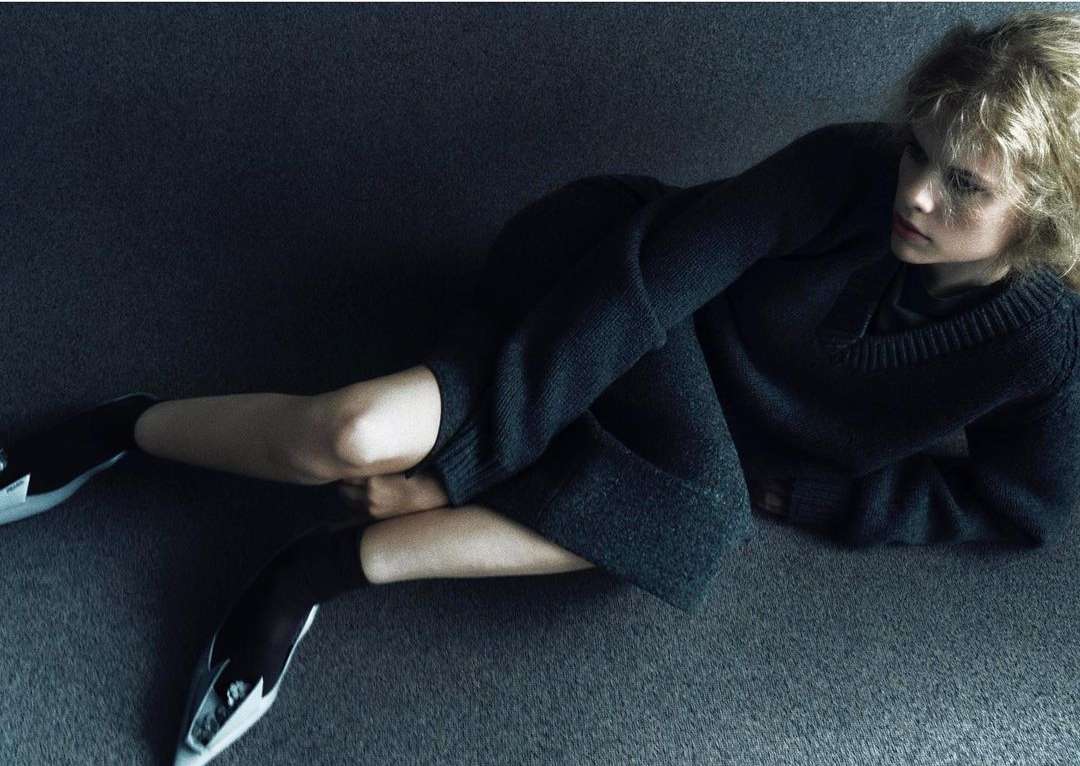 A woman in a dark grey knit sweater and skirt seated on a dark carpeted floor by Hanna Tveite