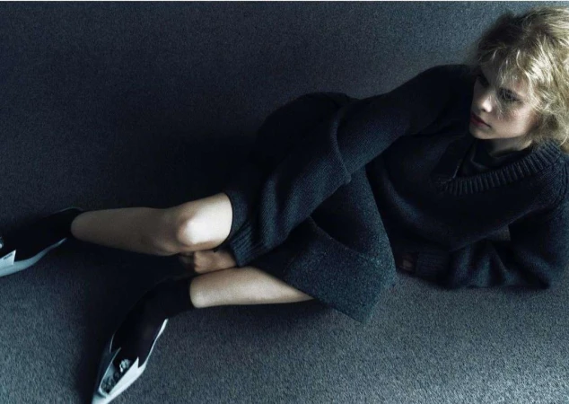 A woman in a dark grey knit sweater and skirt seated on a dark carpeted floor by Hanna Tveite