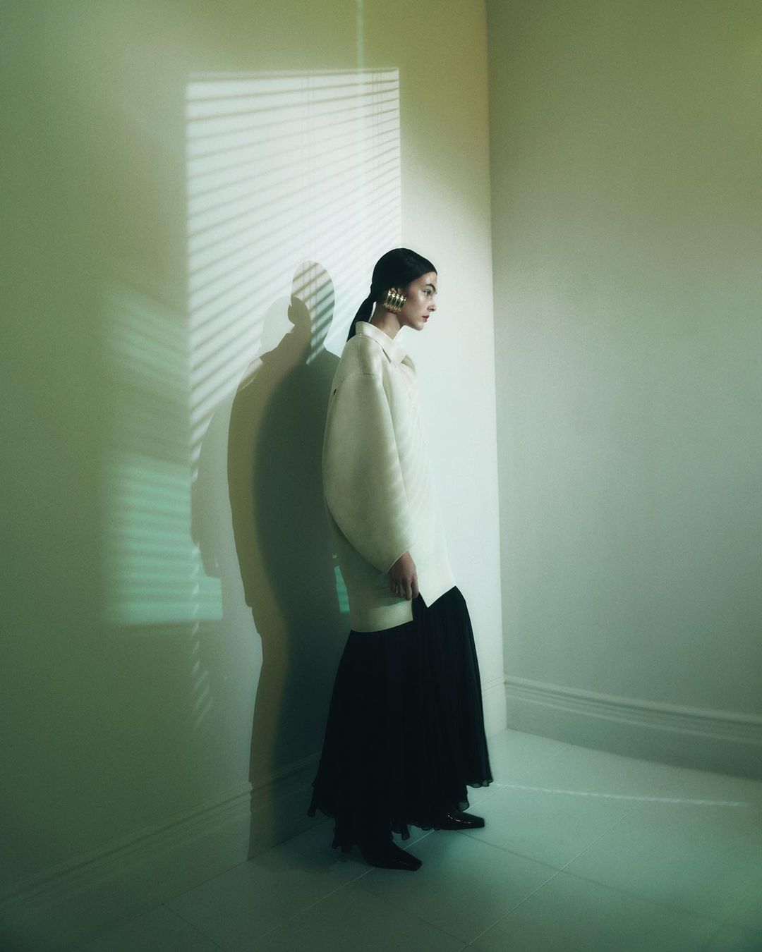 A young woman in a white jacket and black skirt poses in a white room, a shadow from window blinds cast on the wall creating contrast.