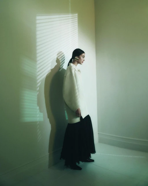 A young woman in a white jacket and black skirt poses in a white room, a shadow from window blinds cast on the wall creating contrast.