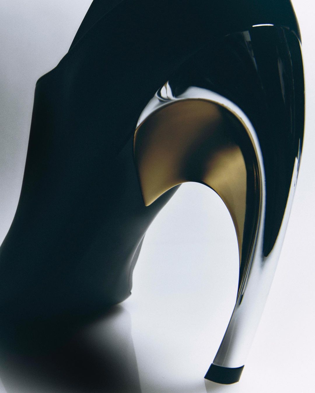 Elegant black high-heel shoe featuring a unique, reflective silver and gold curved heel design, presented on a clean white background.