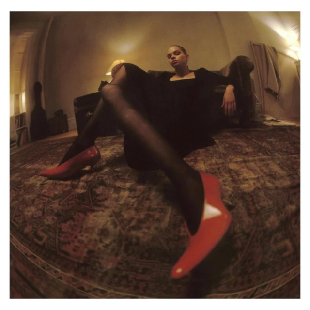 Woman in a black dress and red high heels sits on a couch in a warm-toned room with a patterned carpet. She gazes into the camera.
