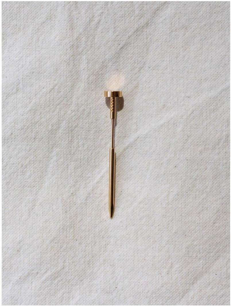Elevated view of a gold-colored collar pin with a round disc head lying on a textured, light-colored fabric surface.