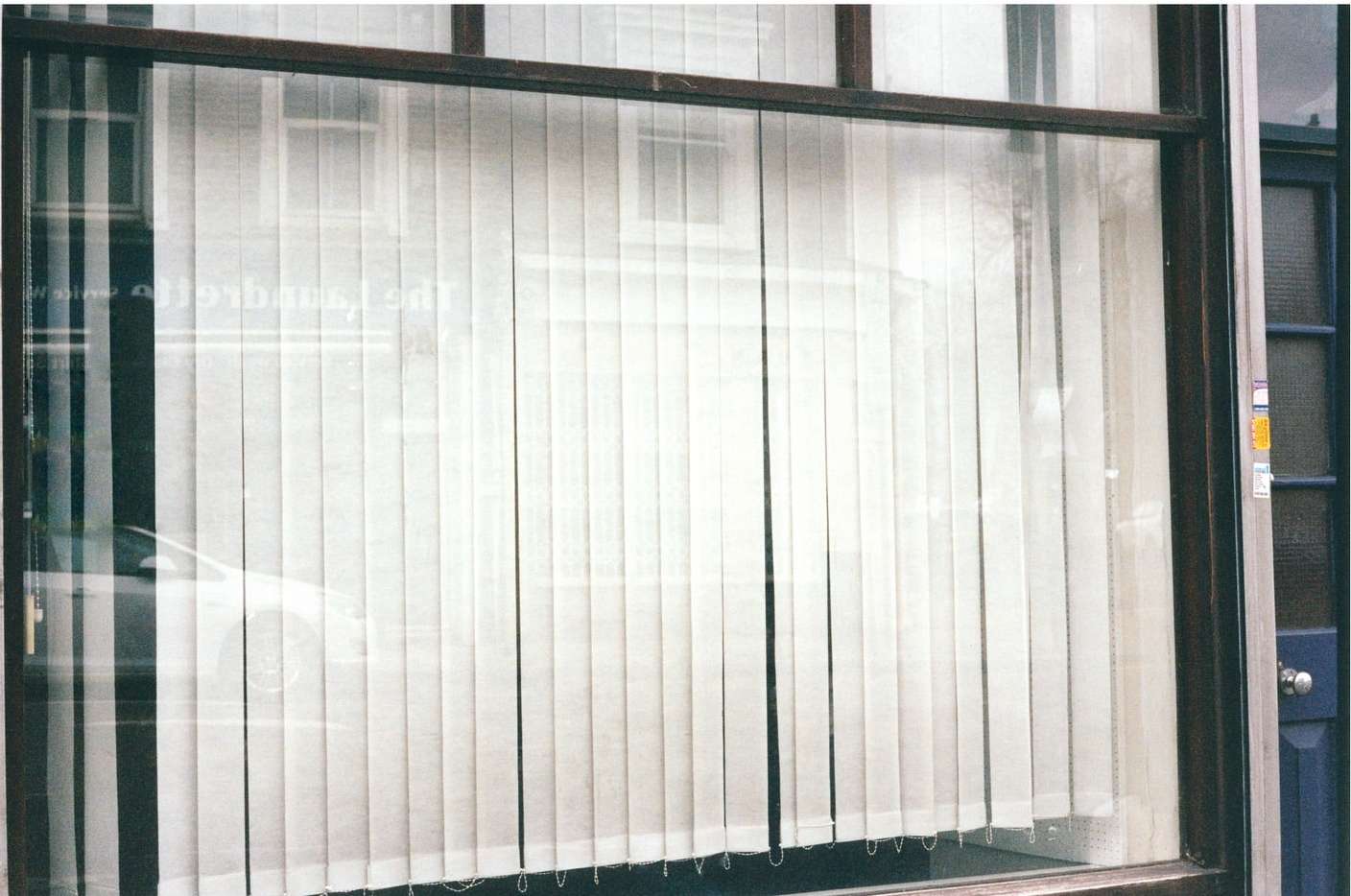 View through a window with vertical blinds, reflecting a street scene and a passing vehicle, creating an urban and architectural scene.
